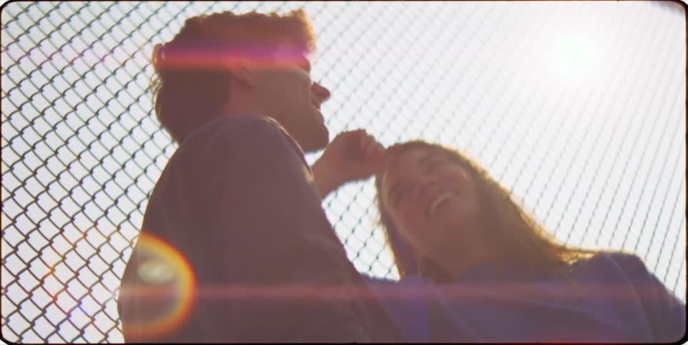 A man and a woman sit on the ground of a fenced pedestrian overpass touching each other and laughing as the sun shines brightly in the background