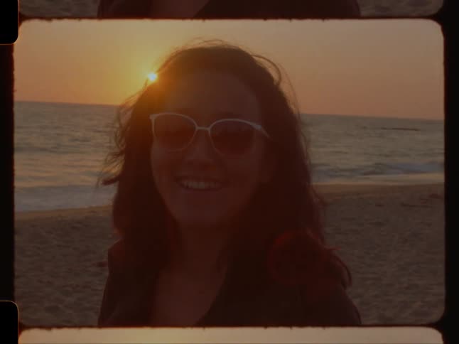 A woman with wavy hair and sunglasses smiles as the sun sets behind her on the beach