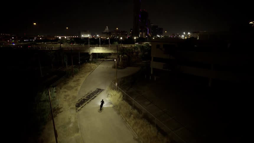 A motorcycle drives down a road at night and the camera ascends revealing a city in the background