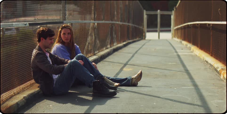 A man and woman sit on the curb of an overpass while the man talks