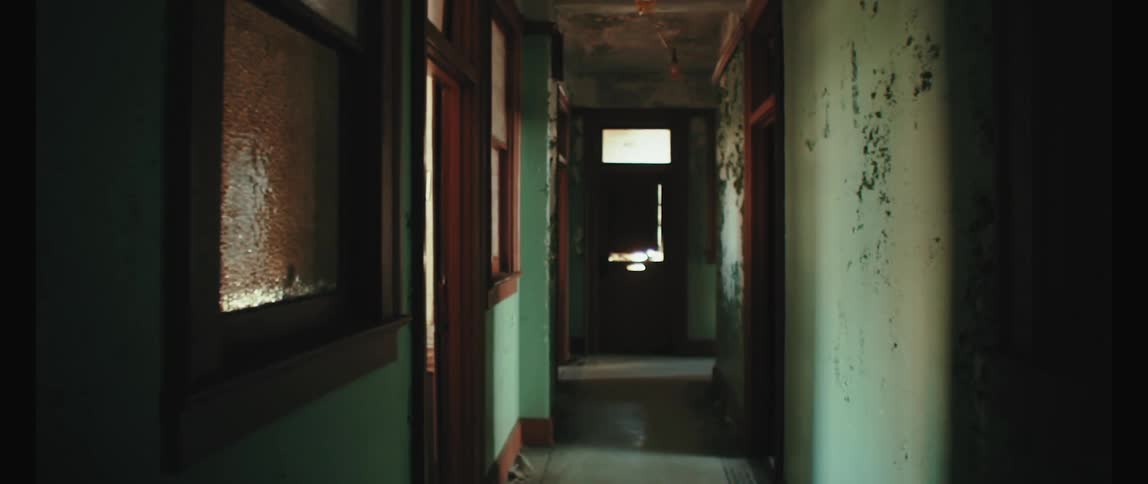 A dark and narrow hallway with peeling paint and wooden door frames leads to a door at the end