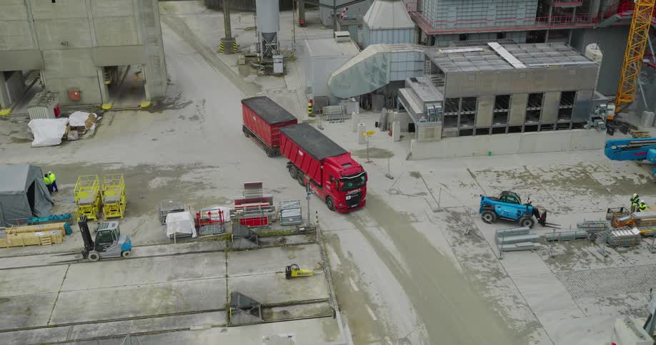 A red truck with two trailers drives through an industrial complex with large concrete structures