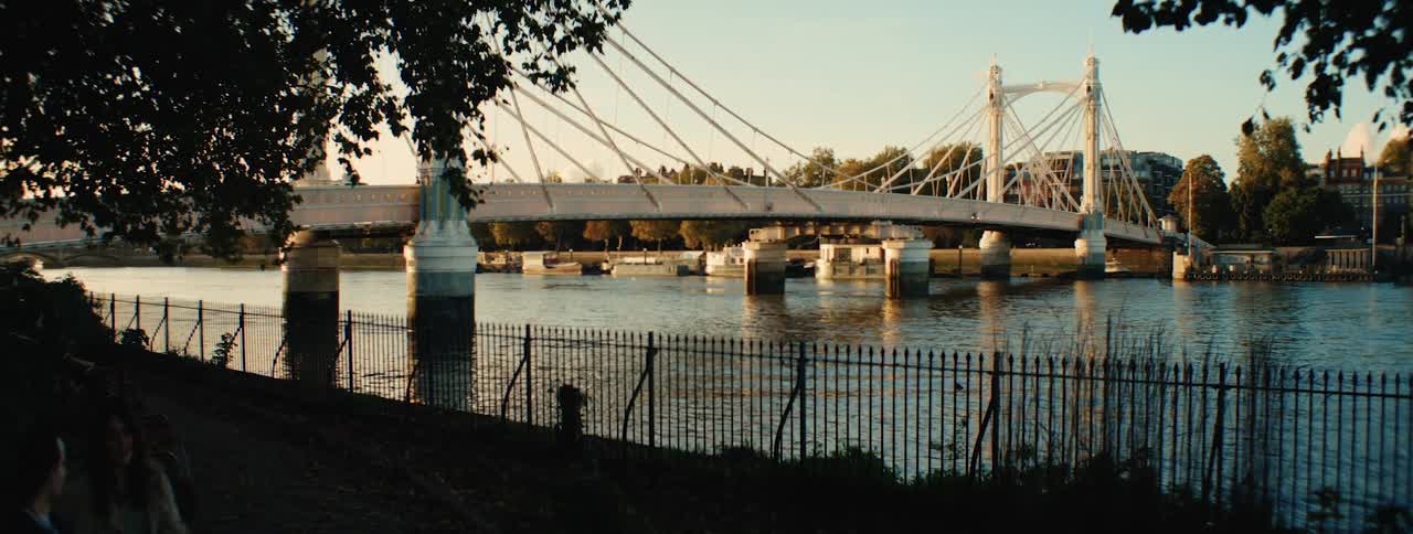 Two people sitting on a bench in a square by a lake with a bridge in front