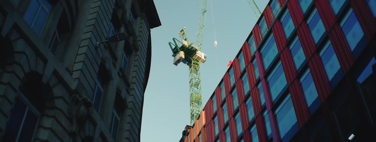 A green construction crane rises between a historic stone building and a modern red and glass structure under a clear sky