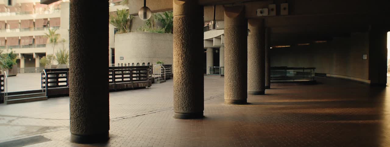 A long narrow corridor in the barbican centre with concrete columns and a tiled floor dimly lit with two people walking at the end