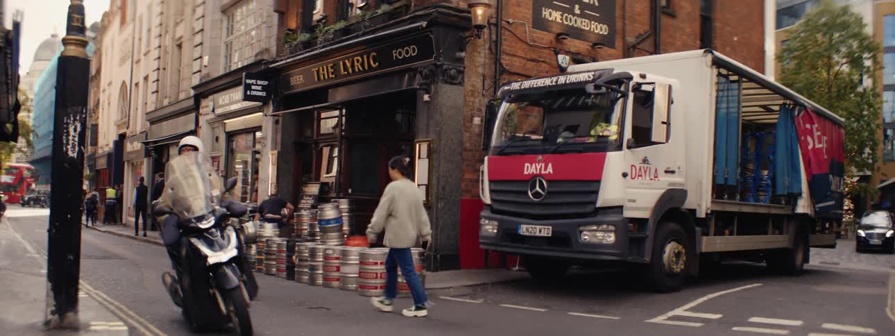 A delivery truck is parked beside a pub on a city street while a person walks across the road and another person works stacking beer kegs
