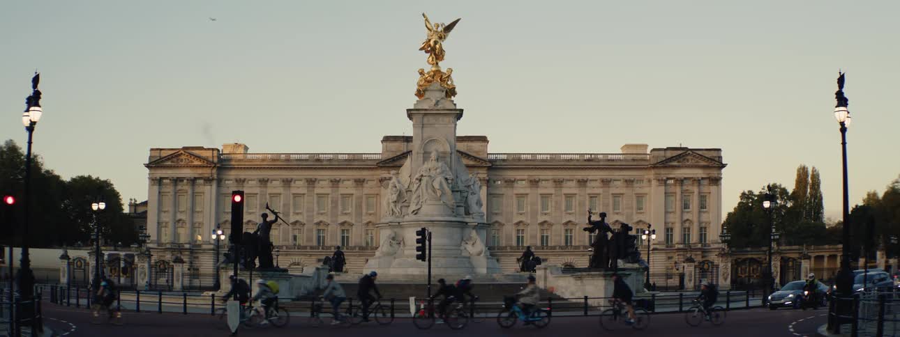 Buckingham palace and the victoria memorial are shown at dusk or dawn with cyclists ride in both directions in the foreground while cars wait at a traffic light