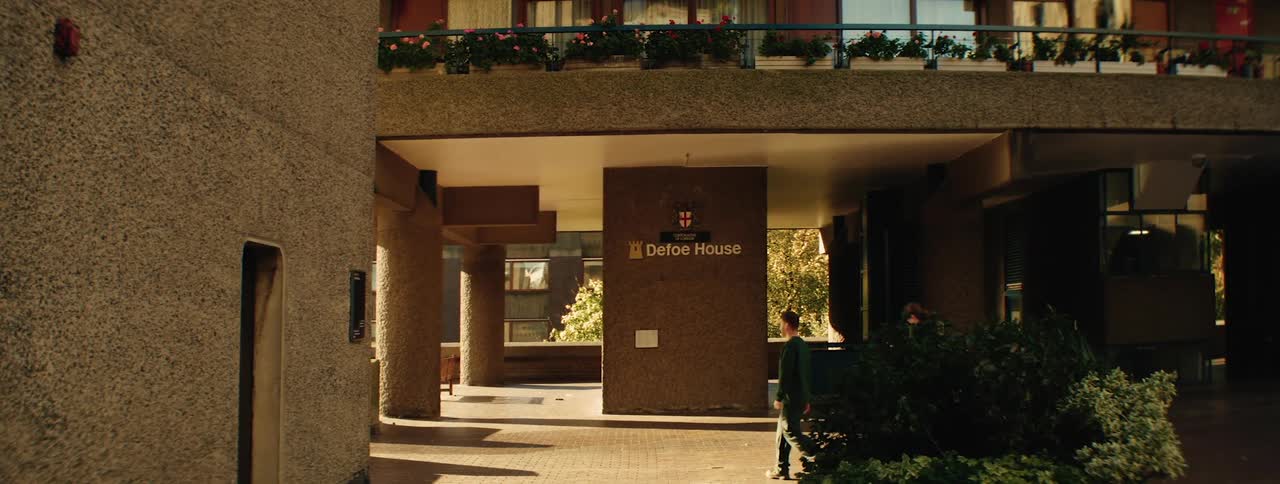 Two people walk past the entrance of a modern building named defoe house featuring large pillars and a balcony adorned with flowers