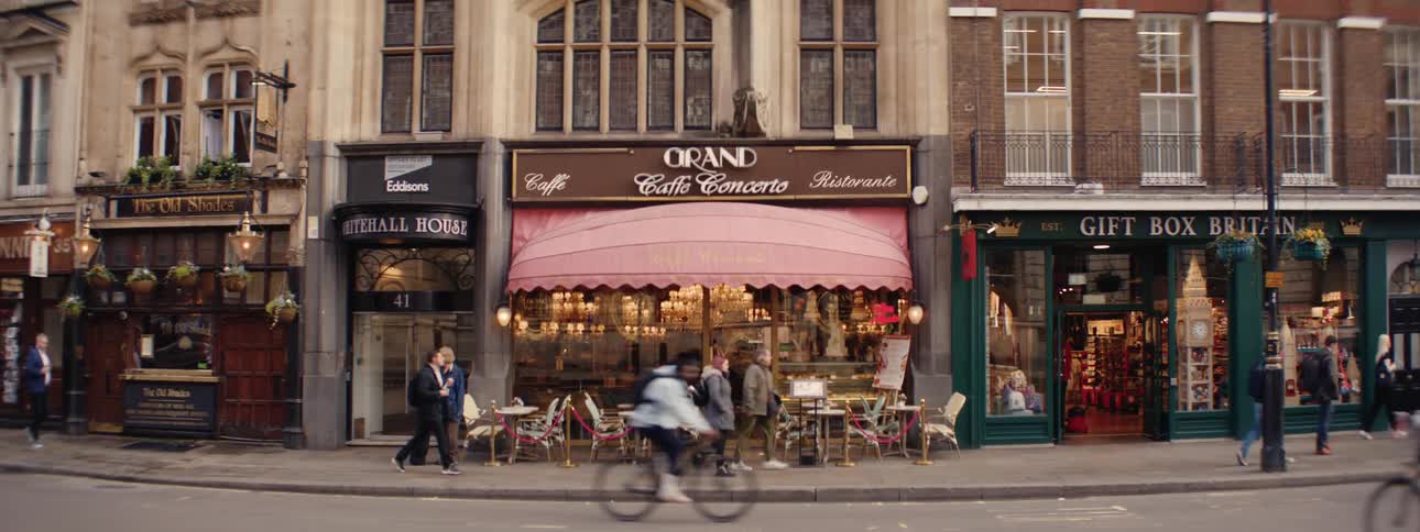 A bustling street scene shows pedestrians walking past a quaint european style cafe with a pink awning flanked by other shops in a city setting