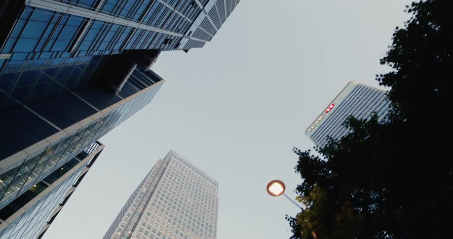 A lowangle shot from street level looks up at the towering skyscrapers of canary wharf including the hsbc building with tree branches and a lit lamppost in the foreground