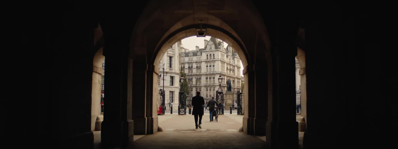Some people passing under a dark corridor with pillars on the sides and in the background the external environment with some buildings