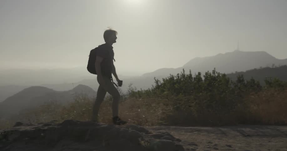 A man with a backpack looking over the edge of a cliff and then walking away