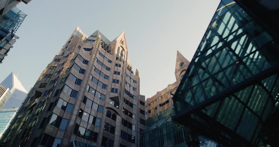A tall angular building with many windows is viewed from below nestled among other modern buildings in a city setting