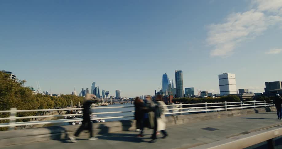 A moving shot from a vehicle on a bridge captures pedestrians in autumn attire walking in both directions on a parallel sidewalk with a river and cityscape in the background