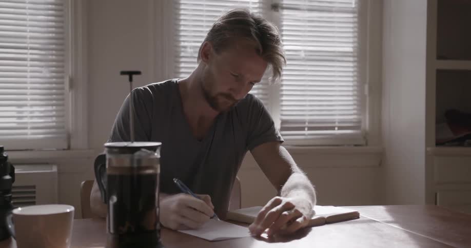 A man sitting at a table in his house writing on a piece of paper