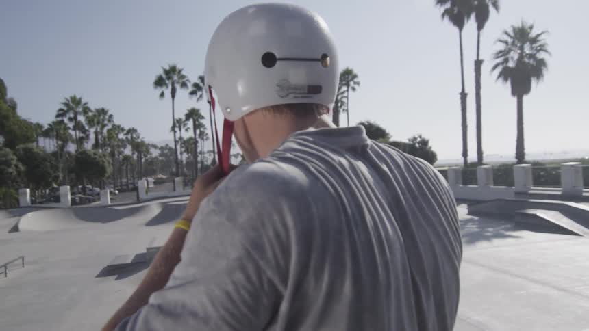 A skateboarder puts on a helmet at a skatepark on a sunny day