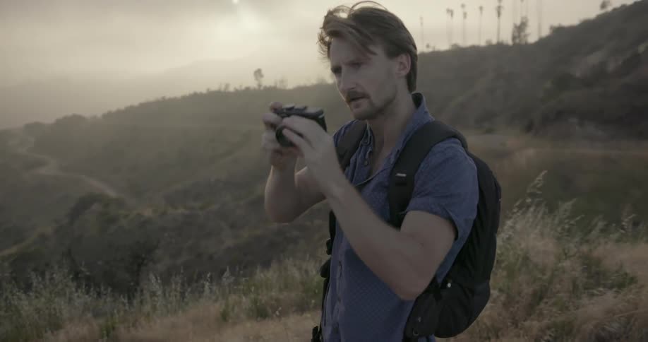 A man wearing a backpack is taking a photo in a hilly area