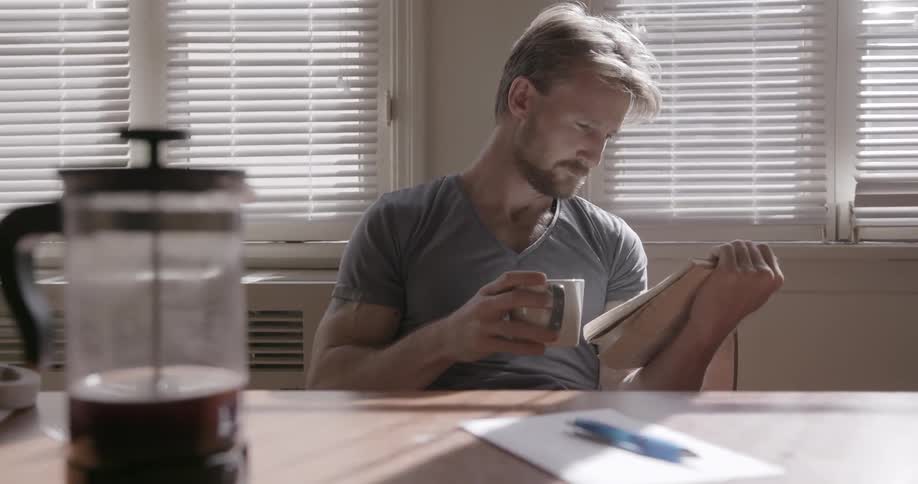 A man sitting at a kitchen table is drinking from a coffee cup and reading