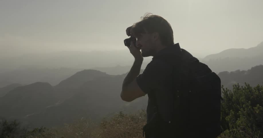 A man wearing a backpack is taking a photo in a hilly area on a hazy day