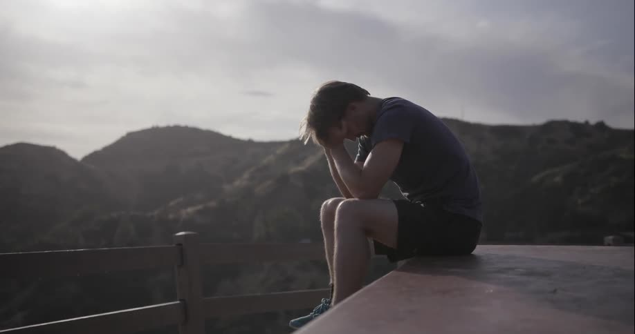A man sitting on a platform at a scenic lookout raises his head from his hands