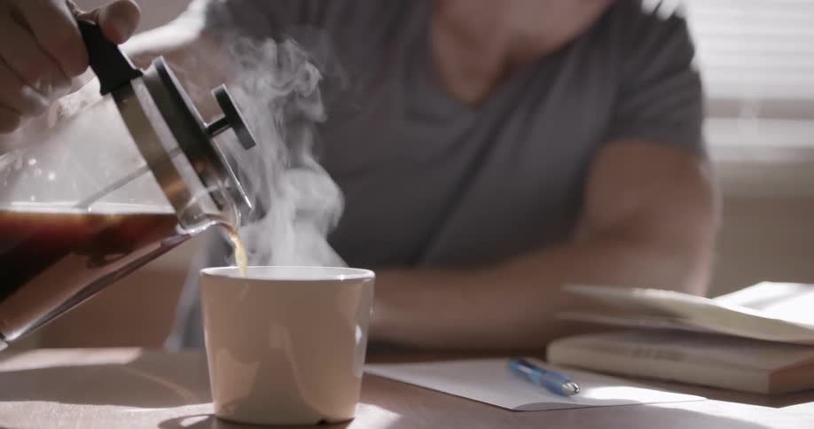 Man pours hot coffee into a mug while sitting at a table