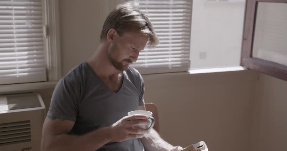 A man is reading a book and drinking coffee at a kitchen table
