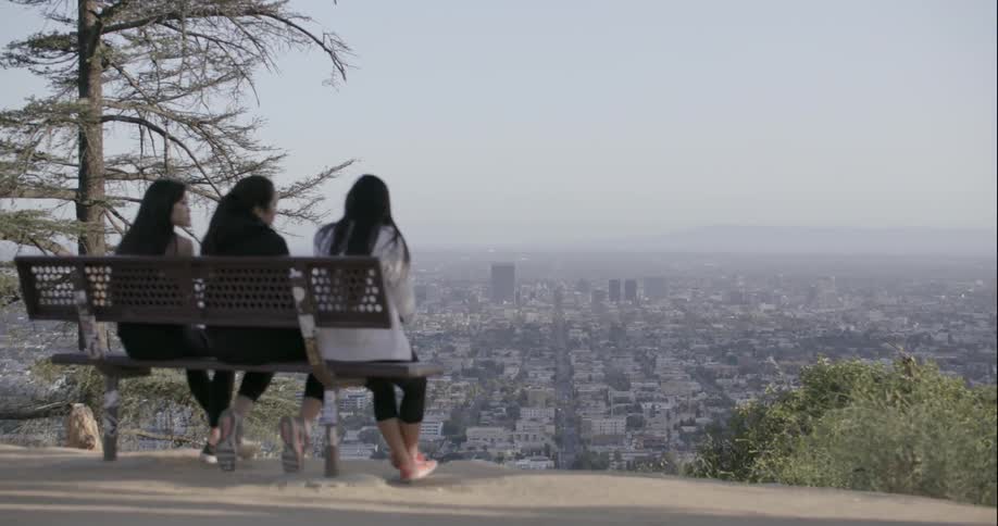 Three women sit on a bench overseeing a city below