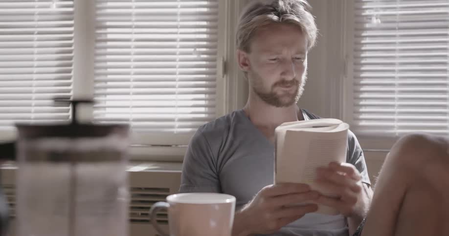 A man is reading a book at a kitchen table during the day