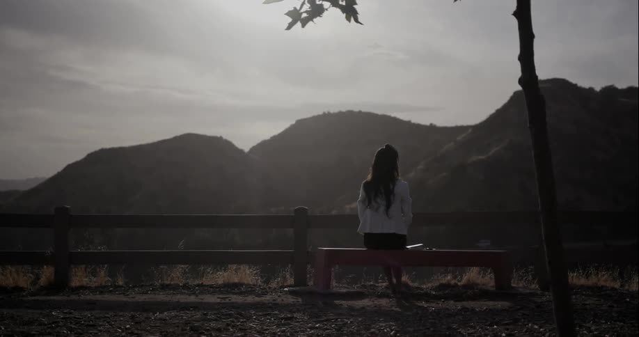 A woman sits on a bench overlooking a hilly area