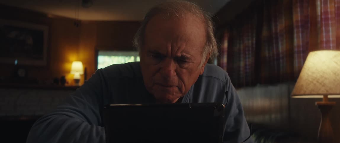 An elderly man sits at a desk takes off his glasses and looks at a tablet screen confused