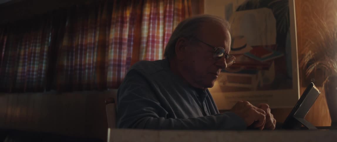 An elderly man is seated at a table intently using a tablet device with plaid curtains