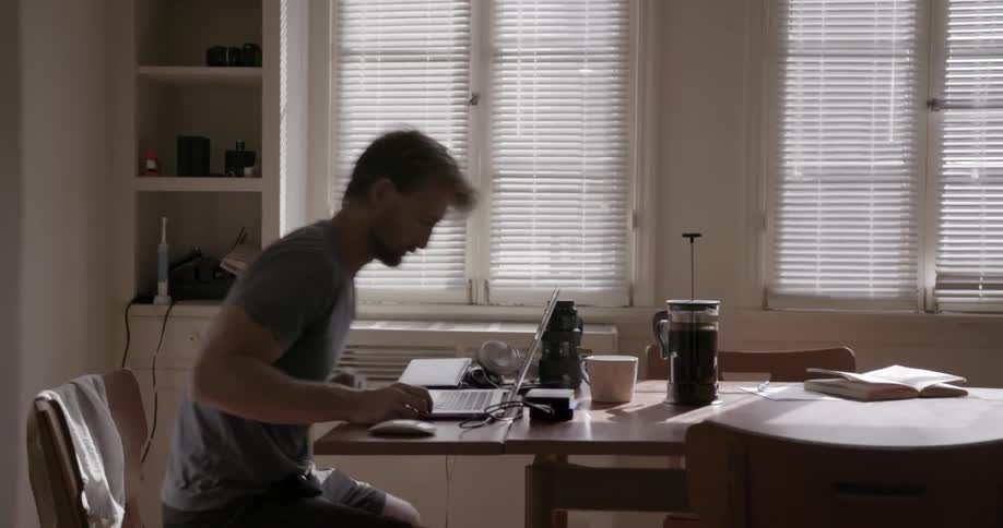A man sits down to work at a laptop sitting on a dining room table