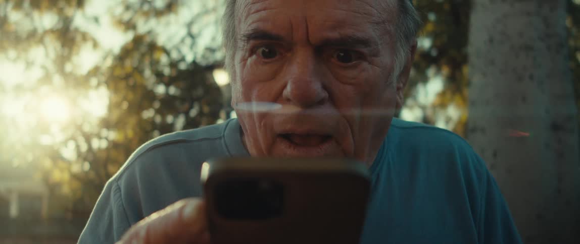 An elderly man with a surprised expression transitioning to a smile as he looks at his cell phone with a bright lens flare obstructing a portion of his face