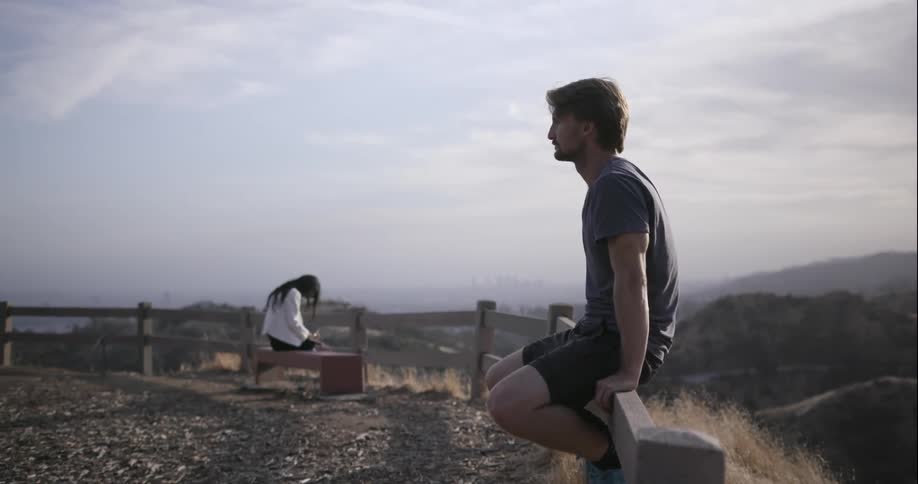 A man is sitting on a ridge fence while a woman is sitting on a bench in the background