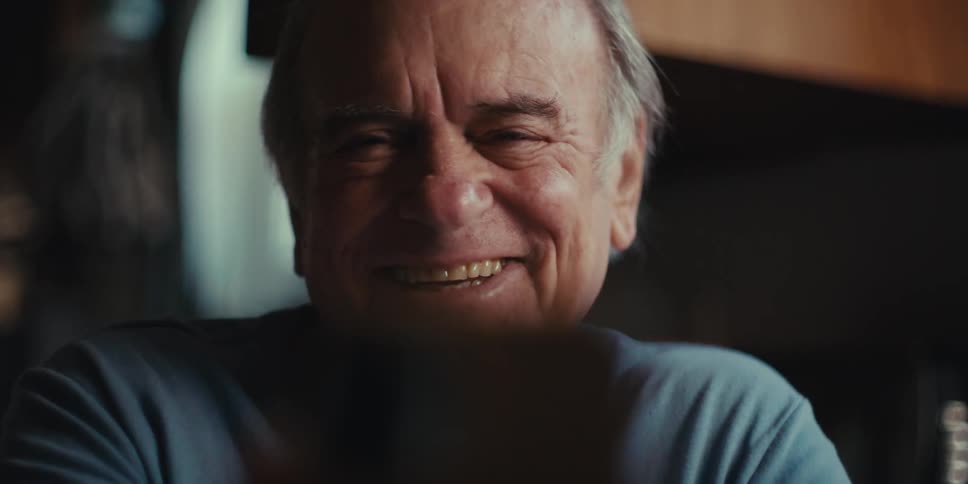 A close up shot of an older man shows him transitioning from a neutral expression to a wide genuine laugh before returning to a neutral expression