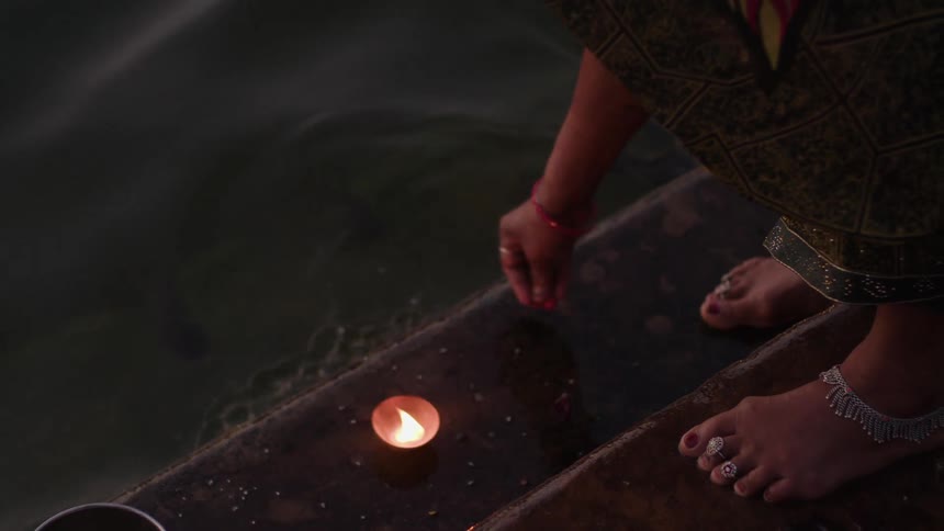 A woman wearing anklets lights a small candle on the edge of a river her bare feet resting on the stone steps