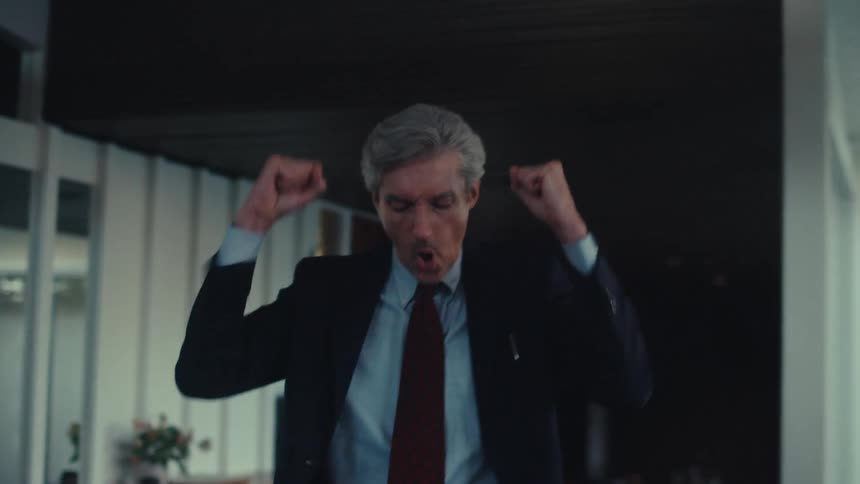 A person in a suit and tie is standing indoors celebrating or expressing triumph