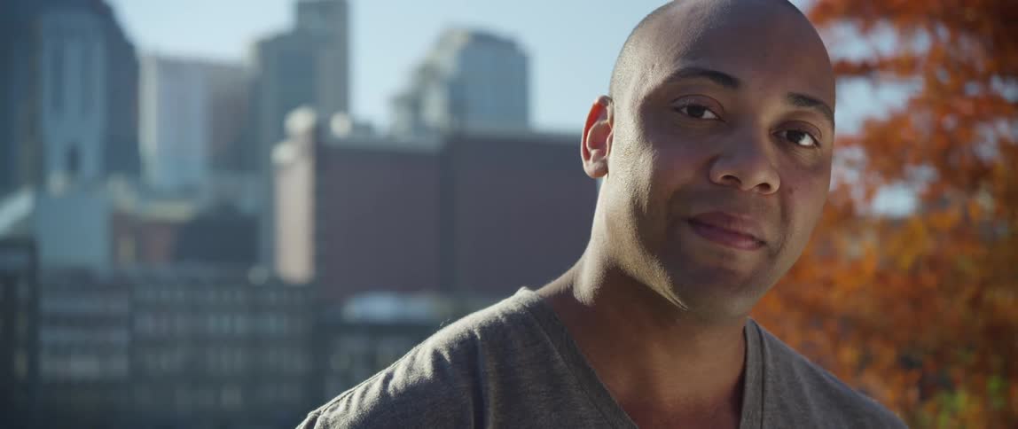 Man smiles on a sunny day with a city behind him