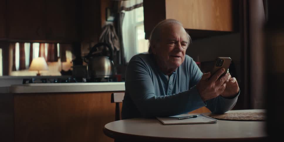 An elderly man smiles and laughs while looking at his phone at home