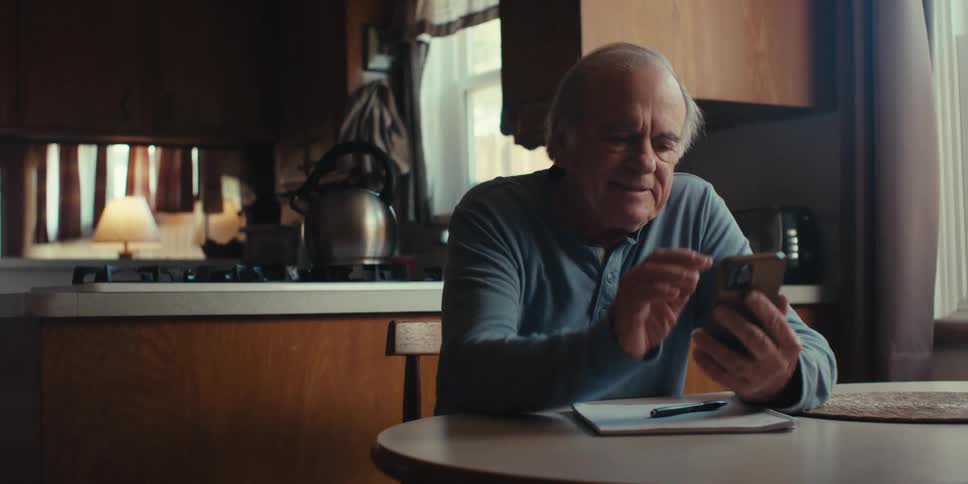 An elderly man sits down at his dining room table and scrolls on his smartphone