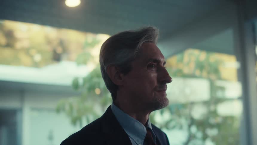 A man in a suit stands looking out a large window with a thoughtful expression