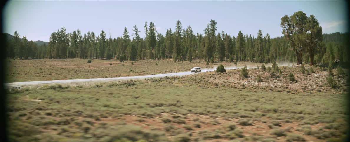 A white suv speeds down a dirt road through a vast open landscape dotted with tall pine trees