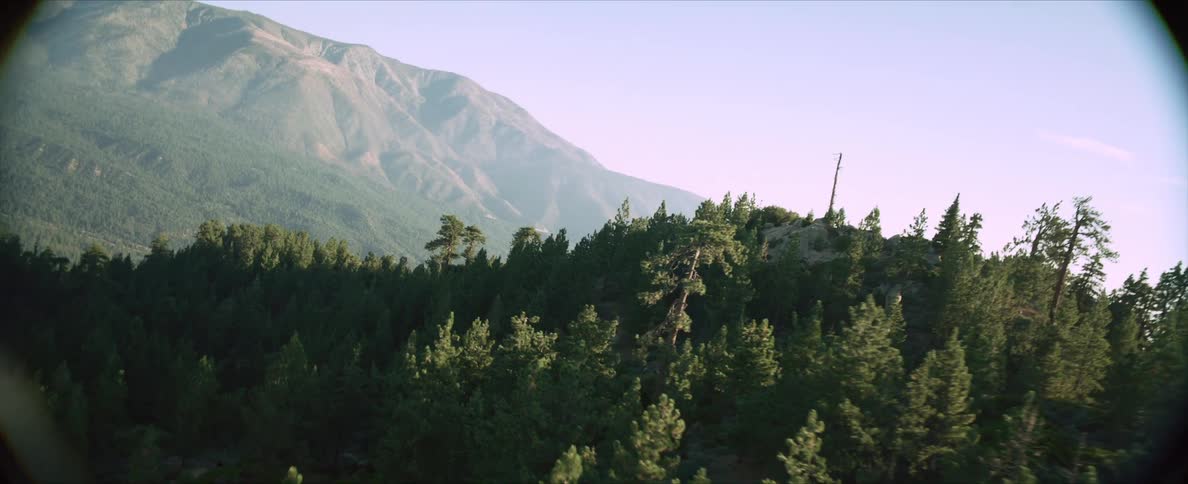 An aerial view reveals a forested mountain slope with a building nestled amongst rocks on the mountaintop