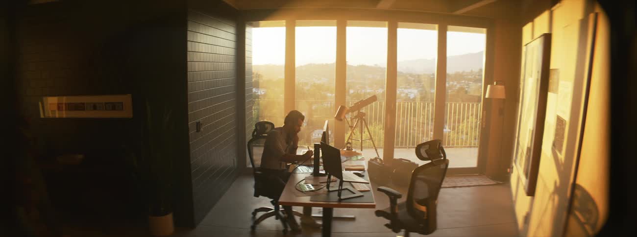 Two people are working at a desk with computers in a room with large windows a telescope and a scenic view of a city and mountains in the background