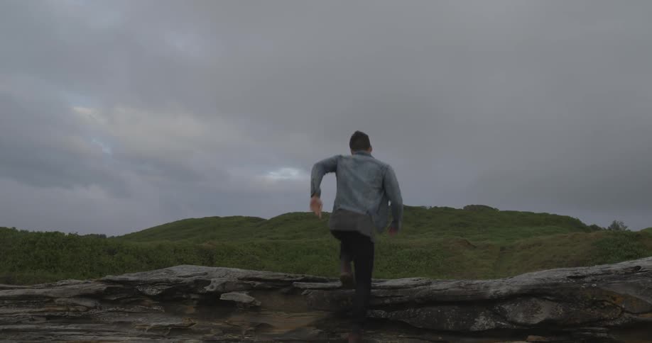 A man runs up rocks near a grass covered hill