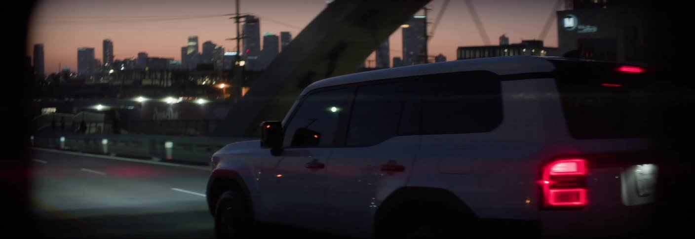 A white suv drives on a bridge at dusk with a city skyline visible in the background