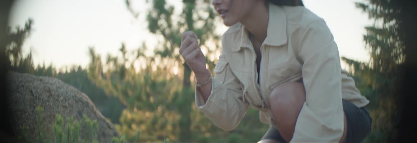 A woman hiking in the wilderness picks up and observes a small wildflower in the morning