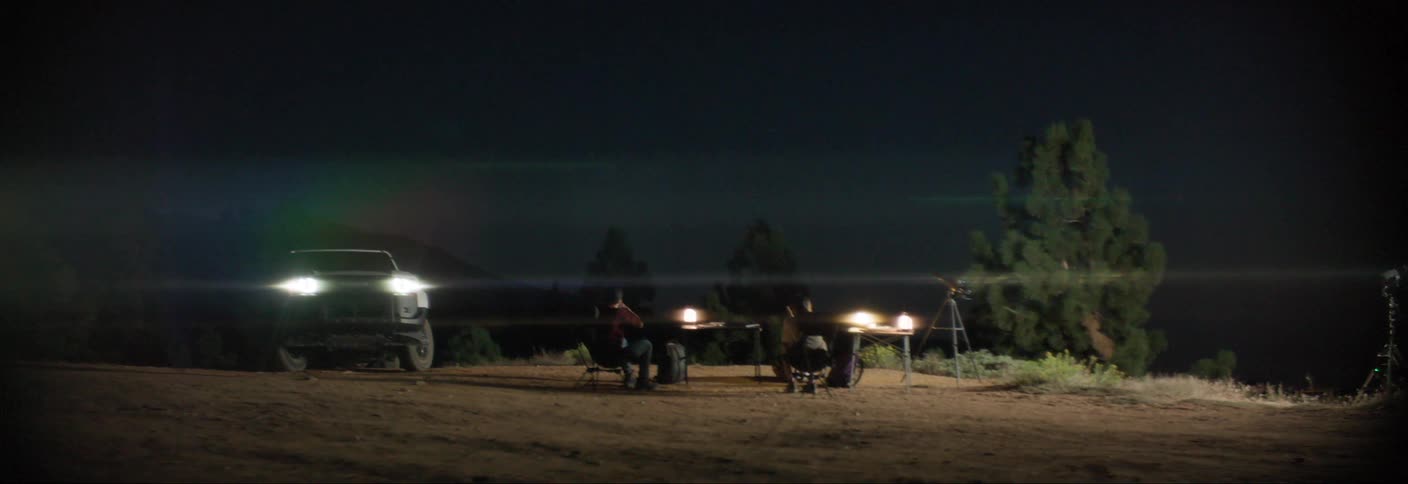 Two people sitting on a chair outdoors at night in a natural setting and a pickup truck next to it with its lights on