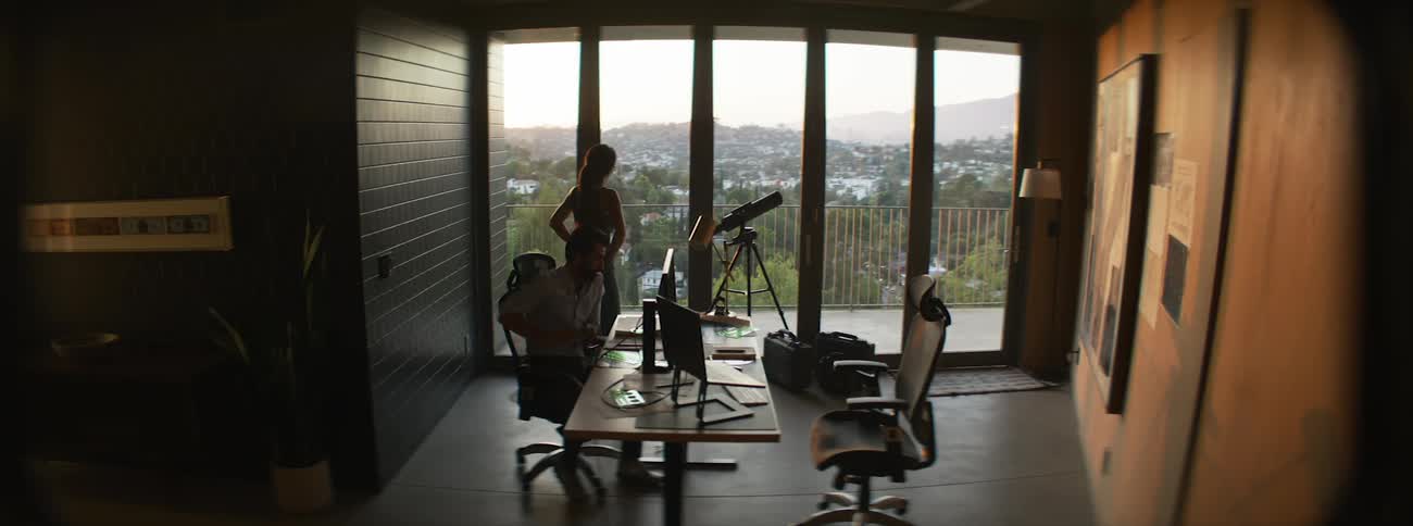 A wide shot shows a man working at his computer and a woman standing by a telescope in a modern home office with a large window overlooking a city at sunset