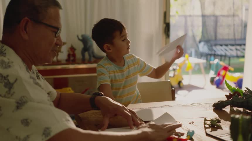 Grandfather sitting at the table making a paper airplane and his grandson playing next to him